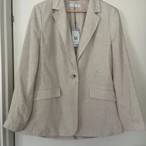 Beige kavaj från Primark i linne - Stilren beige kavaj från Primark i linneblandning. Klassisk modell med en knapp framtill, två fickor med lock och snygg krage. Perfekt för dig som gillar en clean och fräsch look. Helt ny med prislapp kvar, passade inte tillsammans med mina linnebyxor 