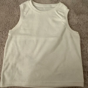 Vit ribbad linnetopp i bomull  - Enkel och stilren vit ribbad linnetopp i 100% bomull. Modellen är ärmlös med rund halsringning och har en mjuk, stretchig känsla. Perfekt att matcha med jeans eller kjol för en clean look. (Storlek L men passar även den som bär M) 