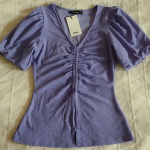 Ljuslila topp med puffärm från Bik Bok - Supersöt ljuslila topp från Bik Bok med korta puffärmar och v-ringning. Toppen har snygg rynkdetalj framtill och är figurnära i modellen. Materialet är mjukt och lite texturerat, perfekt för dig som gillar trendiga detaljer och en feminin look.