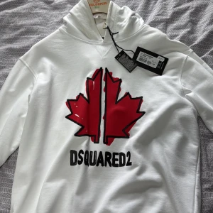 Vit hoodie från Dsquared2 med lönnlöv - Säljer en vit hoodie från Dsquared2 med stor röd lönnlövslogga och svart text på bröstet. Tröjan har huva, långa ärmar och är gjord i mjuk bomull. Perfekt statement-plagg för dig som gillar streetwear och snygga loggor.