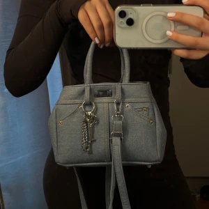 Jeans handväska med kedjedetalj - Snygg handväska i ljusblå jeans med coola fickdetaljer och silverfärgade kedjor och nyckelberlocker. Väskan har både handtag och justerbar axelrem, perfekt för att lyfta din outfit med en trendig denimlook.