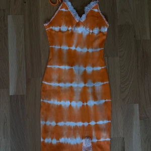 Orange och vit ribbad kortklänning - Superfin ribbad kortklänning i orange och vitt batikmönster. Klänningen har smala axelband, spetsdetaljer vid urringning och nederkant samt en liten slits framtill. Perfekt för sommarens varma dagar och ger en riktigt fräsch vibe.
