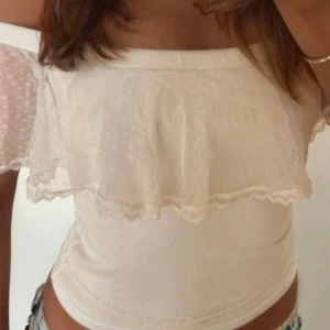 Vit offshoulder topp med volang - Supersöt vit offshoulder topp med volang och spetsdetaljer. Tunt, lätt material med diskret prickigt mönster och romantisk känsla. Perfekt till jeans eller kjol för en fräsch look.