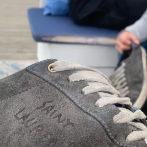 Grå mocka sneakers från Saint Laurent - Snygga grå sneakers från Saint Laurent i mjuk mocka med vita snören och blå detaljer på plösen. Klassisk rund tå och vit sula med fiskbensmönster. Perfekta för dig som vill ha en stilren och lyxig look. Byten kan vara intressanta, pris ej hugget i sten 🪨 