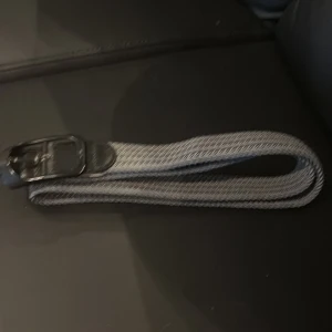 Grått flätat bälte med svart spänne - Snyggt grått bälte med flätad design och svart spänne. Perfekt för att ge outfiten en stilren touch. Bältet är gjort i ett slitstarkt material och passar till både jeans och chinos.