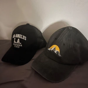 Två svarta kepsar med brodyr 2 för 200 - Säljer två snygga svarta kepsar. Ena har broderad text 'Los Angeles L.A. California' framtill och mesh på sidorna. Den andra har broderat motiv av berg och sol i vitt och gult. Båda har böjd skärm och justerbar passform.