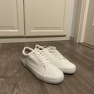 Vita sneakers från Bershka strl 41 - Fräscha vita sneakers från Bershka i storlek 41. Skorna har en clean design med vita snören, rund tå och en brun detalj vid hälen. Tillverkade i syntetmaterial med platt sula, perfekta för en stilren vardagslook.