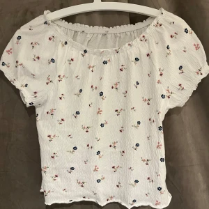 Vit blommig blus - Supersöt vit blus med små blommor i rosa och blått. Blusen har korta puffärmar, resår vid halsringningen och en lätt rynkad struktur i tyget. Perfekt för en somrig look och passar till både jeans och kjol.