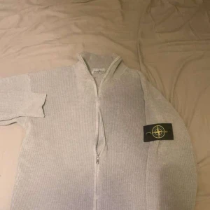 Stone Island kofta - Säljer en ljusgrå stickad kofta från Stone Island med dragkedja framtill och klassisk logopatch på ärmen. Koftan har ribbad struktur, hög krage och långa ärmar. Perfekt för dig som gillar stilrena och exklusiva plagg.