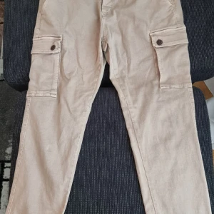 Beige Replay Hyperflex cargobyxor Jaan w34,32 - Snygga beige cargobyxor från Replay Hyperflex med stretchigt material och stora fickor på sidorna. Byxorna har raka ben, knappstängning och klassisk cargostil. Perfekta för dig som vill ha både stil och funktion i ett par byxor.Helt nya med kvitto 