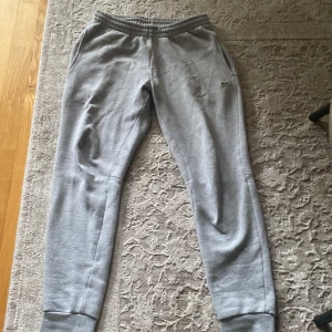Grå mjukisbyxor från Lacoste XS - Slim fit grå mjukisbyxor från Lacoste med broderad krokodil-logga på benet. Byxorna har resår och snörning i midjan samt muddar vid bensluten. Tillverkade i mjukt bomullsmaterial för maximal komfort. Den är XS i män storlek.