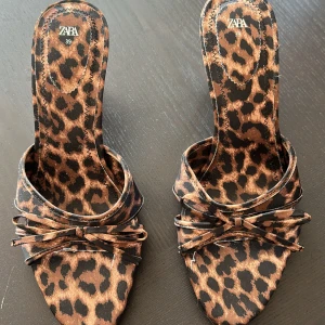 Leopardmönstrade klackar från Zara - Snygga sandaler från Zara med leopardmönster i bruna och svarta toner. Skorna har öppen tå, smal hög klack och en dekorativ rosett framtill. Perfekta för dig som vill sticka ut med ett djurmönstrat statement.
