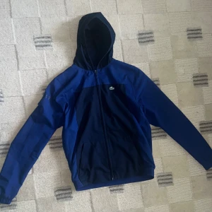 Lacoste vindjacka - Vindjacka från Lacoste Sport. Köpt på NK i mars 2024 för 1900 kr. Mycket fint skick. Storlek 16Y/176 cm - ungefär som en medium i storlek. 