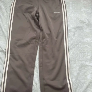 Bruna Adidas byxor med vita ränder - Säljer ett par bruna Adidas byxor med klassiska vita ränder längs sidorna och Adidas-logga på höften. Byxorna har rak passform, resår i midjan och är tillverkade i ett mjukt syntetmaterial. Perfekta för en sportig och avslappnad stil.