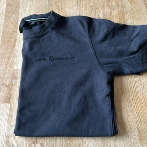 Svart t-shirt från Sail Racing - Svart t-shirt från Sail Racing med diskret logga på bröstet. Klassisk passform och rund halsringning. Tillverkad i mjuk bomull som känns skön mot huden. Perfekt för dig som gillar stilrena och sportiga plagg.