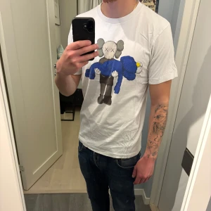 KAWS x Uniqlo vit t-shirt med tryck - Vit t-shirt från Uniqlo i samarbete med KAWS. Framsidan har ett stort färgglatt tryck med KAWS ikoniska figur som bär en blå karaktär. 