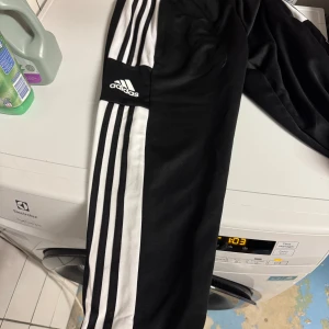 Svarta Adidas mjukisbyxor med ränder - Svarta mjukisbyxor från Adidas med klassiska vita ränder längs sidorna och Adidas-logga på benet. Byxorna har en sportig look och är tillverkade i mjukt material som passar perfekt för chill eller träning. Nya med lapp.