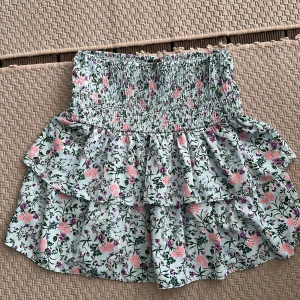 Blommig volangkjol med smock S - Supersöt kort kjol med blommigt mönster i rosa, lila och grönt på ljus botten. Kjolen har bred smock i midjan och dubbla volanger som ger en flowy känsla. Perfekt för dig som gillar romantisk och lekfull stil. Storlek S från Vero Moda
