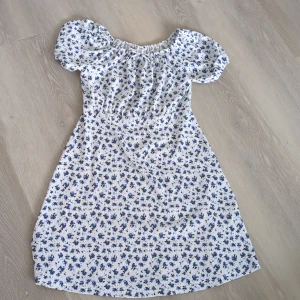 Blommig kortklänning från Zara - Supersöt vit kortklänning från Zara med blått blommönster och puffärmar. Klänningen har smockad resår upptill, liten rosett vid halsen och är tillverkad i ett lätt bomullstyg. Perfekt för sommarens alla tillfällen!