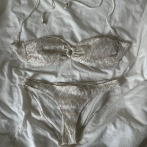 Vit & beige bikini med glittrig guld från H&M - Supersnygg bikini från H&M i glittrigt guld med diskret mönster och snygg ringdetalj framtill på överdelen. Bikinin har bandeau-topp med knytband och matchande trosa. Perfekt för strandhäng och poolparty! ÖVERDELEN ÄR I STORLEK 42 OCH UNDERDELEN I STORLEK 38