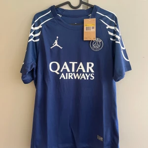 PSG x Jordan blå matchtröja - Snygg blå PSG fotbollströja från samarbetet med Jordan. Tröjan har vita detaljer, klubbmärke på bröstet och Qatar Airways-tryck framtill. Tillverkad i lätt och ventilerande material, perfekt för träning eller match. Unik design med vita linjer på axlarna.