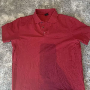 Röd pikétröja från BOSS - Säljer en röd pikétröja från BOSS med klassisk krage och tre knappar framtill. Tröjan är i bomull och har korta ärmar. Diskret BOSS-logga broderad på bröstet. Perfekt för en clean och stilren look.