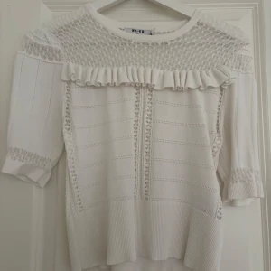 Vit blus med spets och volang från NA-KD - Superfin vit blus från NA-KD med spetsdetaljer upptill, volang längs bröstet och halvärmade ärmar. Blusen har ribbade muddar och ett diskret hålmönster som ger en romantisk vibe. Perfekt till jeans eller kjol för en söt look.