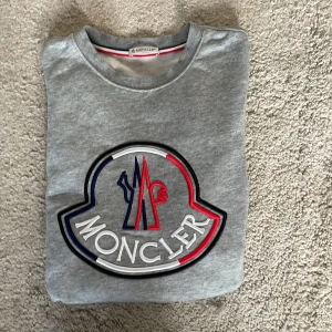 Grå Moncler sweatshirt med stor logga - Snygg grå sweatshirt från Moncler med stor broderad logga i vitt, rött och marinblått på bröstet. Tröjan har rund hals, långa ärmar och ribbade muddar. Perfekt för dig som gillar streetwear och vill sticka ut med ett exklusivt märke.