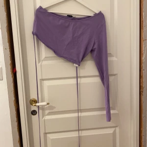 Lila oneshoulder topp från NA-KD - Säljer en lila croppad topp från NA-KD med oneshoulder-design och lång ärm. Toppen har smala knytband i sidorna som ger en snygg detalj och kan justeras. Perfekt för dig som gillar trendiga och unika plagg med cool passform. Pris kan diskuteras!