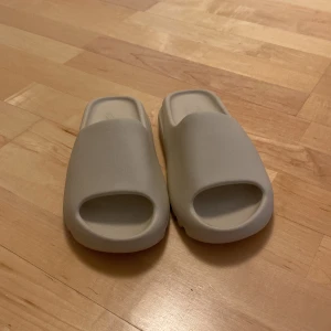 Beige Adidas Yeezy Slide sandaler - Säljer ett par stilrena Adidas Yeezy Slide sandaler i beige. De har chunky, räfflad sula och öppen tå för en modern vibe. Tillverkade i mjuk syntet för maximal komfort, perfekta för sommaren eller chill dagar.