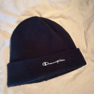 Svart mössa från Champion - En stilren svart mössa från Champion med broderad logga framtill. Klassisk ribbstickad design med uppvikt kant, perfekt för att hålla värmen och ge en clean look. Passar till de flesta outfits och är enkel att matcha.