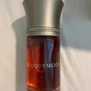 Bloody Wood Eau de Parfum 100ml - Bloody Wood Eau Sanguine från Liquides Imaginaires är en unik parfym i en lyxig, cylindrisk glasflaska med röd vätska och en elegant silverfärgad metallkork. Kommer i en stilren svart box med märkets logga och detaljerad etikett.