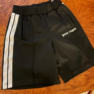 Palm Angels svarta shorts med ränder - (ÄKTA )Svarta Palm Angels shorts med vita dubbla sidoremsor och broderad logga på vänster ben. Elastisk midja och två fickor framtill. Sportig vibe och tillverkade i 100% polyester för en skön och lätt känsla.