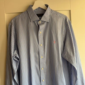 Blåvit randig skjorta Polo Ralph Lauren - Klassisk blå och vit randig skjorta från Polo Ralph Lauren med broderad rosa logga på bröstet. Skjortan har lång ärm, knappar framtill och en tidlös krage. Tillverkad i mjuk bomull och passar perfekt till en clean och stilren look. Stor i storleken. 