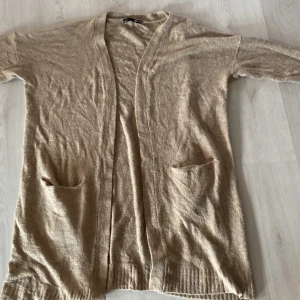 Beige lång kofta från 157 XS/S - Lång, mysig beige kofta från 157 i stickat material. Koftan har två stora fickor framtill, ribbade muddar och en öppen front utan knappar. Perfekt att slänga på sig över en t-shirt eller hoodie för en chill look.