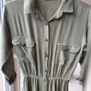 Olivgrön långklänning med knappar - Olivgrön långklänning med skjortkrage, knappar hela vägen framtill och två bröstfickor med lock. Klänningen har långa ärmar som kan vikas upp och fästas med knapp. Materialet är mjukt och faller snyggt.
