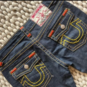 True Religion jeans med kontrastsömmar - Mörkblå jeans från True Religion med ikoniska gula och orange kontrastsömmar, stora bakfickor med broderad logga och detaljerade nitar. Klassisk femficksmodell i jeansmaterial med raka ben och låg midja. Perfekta för dig som gillar streetwear och vill sticka ut. Dom är dock avklippta! Storlek 29
