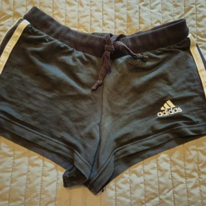 Svarta Adidas shorts med snörning - Svarta sportiga shorts från Adidas med vit logga och vita ränder på sidorna. Mjuk resår i midjan med snörning för perfekt passform. Perfekta för träning eller chill. Klassisk och enkel design som alltid funkar.