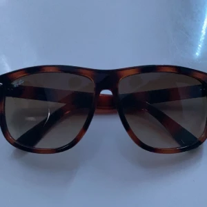 Ray-Ban bruna Wayfarer solglasögon - Snygga Ray-Ban Wayfarer solglasögon i brunmelerad plast med klassisk fyrkantig form. Glasen är tonade och bågarna har Ray-Ban-logga i silver på sidorna. Perfekt accessoar för att lyfta din stil i solen.