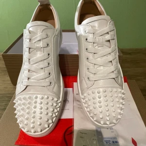 Vita Louboutin sneakers med nitar - Snygga vita sneakers från Christian Louboutin i skinn med ikonisk röd sula och coola nitar på tån. Skorna har snörning och en stilren, modern look som sticker ut. Perfekta för dig som vill ha något extra och vågar sticka ut i mängden. kvitto medföljer!