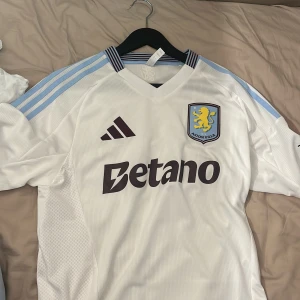 Aston Villa vit bortatröja Adidas M - Aston Villa bortatröja i vitt från Adidas, storlek M. Tröjan har ljusblå ränder på axlarna, klubbmärke på bröstet och sponsortryck. Tillverkad i ventilerande polyester med ribbade detaljer vid kragen. Perfekt för dig som vill visa ditt stöd för Villa. Köptes i vintras på jd sports i London. Aldrig använd bara hängt på min tröj ställning. Storlek M