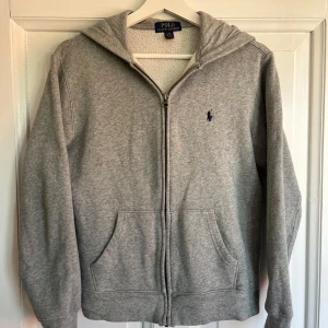 Grå hoodie från Polo Ralph Lauren - Klassisk grå hoodie från Polo Ralph Lauren med dragkedja, huva och två fickor framtill. Broderad marinblå logga på bröstet. Perfekt för chill dagar och enkel att matcha med jeans. Hoodien har storlek 14-16/L i barnstorlekar, tröjan har egentligen storlek S. Hoodien har en mindre defekt som ni kan se på bilden, bortser från det så är Hoodien fläckfri. Tveka inte att fråga om ni vill ha fler bilder eller undrar något!