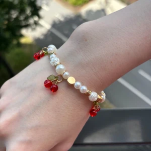 Pärlarmband med körsbärsberlocker - Supergulligt armband med vita pärlor, små rosformade pärlor och guldfärgade detaljer. Hängande körsbärsberlocker i rött och grönt ger en lekfull vibe. Perfekt accessoar för dig som vill sticka ut med något unikt och färgglatt.