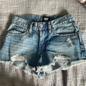 Blå jeansshorts med slitningar  - Snygga blå jeansshorts från lager 157 i storlek XXS. De är lågmidjade med råa, fransiga kanter och slitna detaljer framtill för en cool, avslappnad vibe. Klassisk femficksmodell med knapp och dragkedja. Perfekta för sommardagar och festivalhäng. Säljer då de inte kommit till användning! Använda en gång 