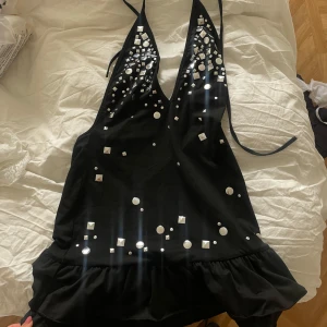 Svart kort klänning med paljetter - Supersnygg svart kort klänning med djup v-ringning och volang nertill. Klänningen har glittriga paljettdetaljer framtill och knytband i nacken. 