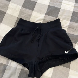 Svarta Nike shorts med snörning - Svarta shorts från Nike med bred resår i midjan och snörning framtill. Klassisk vit Nike-logga på ena benet. Mjukt material, perfekt för träning eller chill. Modellen är kort och har en loose fit som ger en avslappnad vibe.