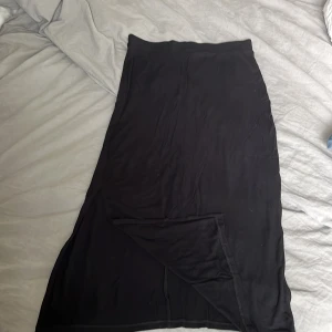 Svart långkjol från Indiska - En svart långkjol från Indiska i mjukt och stretchigt material. Kjolen har en enkel och clean design utan mönster, med rak passform och resår i midjan. Perfekt för dig som gillar minimalistisk stil och vill ha något bekvämt och snyggt.