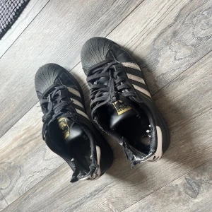 Svarta Adidas Superstar sneakers - Klassiska svarta Adidas Superstar med vita ränder och gummitå. Skorna har snörning, gulddetaljer på plösen och är tillverkade i skinn. Den ikoniska designen med rund tå och platt sula gör dem till en tidlös streetwear-favorit.