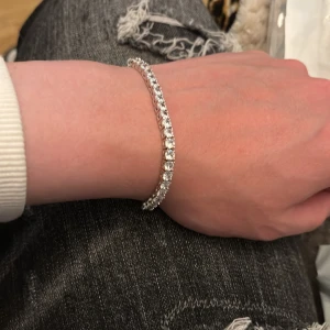 Glittrigt silver tennisarmband - Snyggt tennisarmband i silverfärg med gnistrande stenar runt hela bandet. Armbandet har en klassisk och stilren design med rundade stenar som ger en lyxig känsla. Perfekt för dig som vill ha något som verkligen glittrar och drar blickarna till sig.