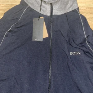 Svart och grå kofta från BOSS - Snygg navy blue  och grå kofta från BOSS med broderad logga på bröstet. Koftan har hel dragkedja, höga muddar och krage i kontrasterande grått. Perfekt för en clean och sportig look. Materialet är mjukt och känns skönt mot huden.